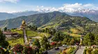 A panoramic view of South Sikkim featuring the golden Buddha Park of Ravangla set against lush green hills and terraced farms, with the snow-capped Himalayan peaks visible in the distant background.